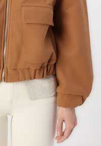 Veste bomber en polaire marron avec fermeture éclair, ourlet plissé et poche latérale. Portée sur un haut gris clair et un pantalon crème. Texture douce.