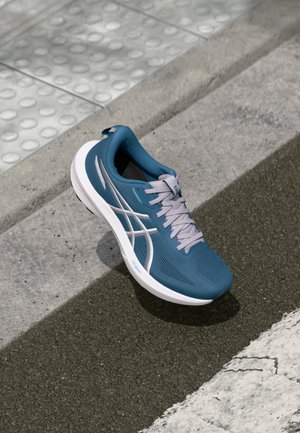 Chaussure de sport bleue avec une tige texturée, accents blancs, semelle flexible et lacets gris. Positionnée sur une surface en béton texturé.