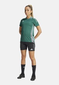 Maglietta sportiva verde con accenti grigi, pantaloni corti neri, calze nere e scarpe sportive. Tre strisce bianche sulla maglietta e sui pantaloni.