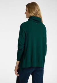 Maglione verde scuro a maniche lunghe con colletto alto e arrotolato. Tessuto morbido, vestibilità comoda e spacchi laterali. Dettaglio del logo piccolo sul retro.