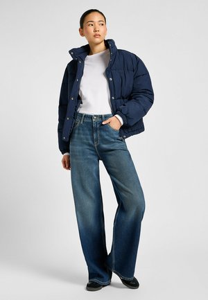 Marineblå puffjakke med høy krage, kombinert med vide blå jeans. Myk tekstur, metallklipsknapper og avslappet passform. Svarte fottøy synlige.