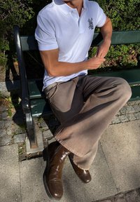 Personne vêtue d'un polo blanc, d'un pantalon marron à jambes larges et de bottes marron foncé, assise jambes croisées sur un banc vert dans un parc en plein air.