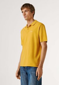 Homme portant un polo à manches courtes jaune moutarde et un jean bleu, debout avec une expression neutre devant un fond clair uni.