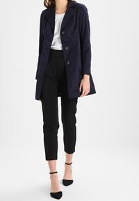 Cappotto trench doppiopetto blu navy con colletto a scialle e maniche lunghe; abbinato a pantaloni neri a vita alta e décolleté a punta.
