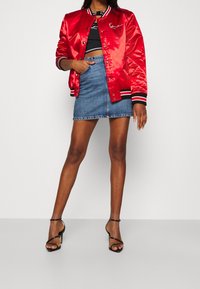 Röd satin bomberjacka, kort svart topp, denim minikjol och svarta klackar med remmar. Jackan har svarta och vita randiga detaljer.