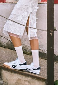 White Reebok sneakers with black accents, paired with white ribbed socks, and a white sheer floral dress with slits, on a stone surface.