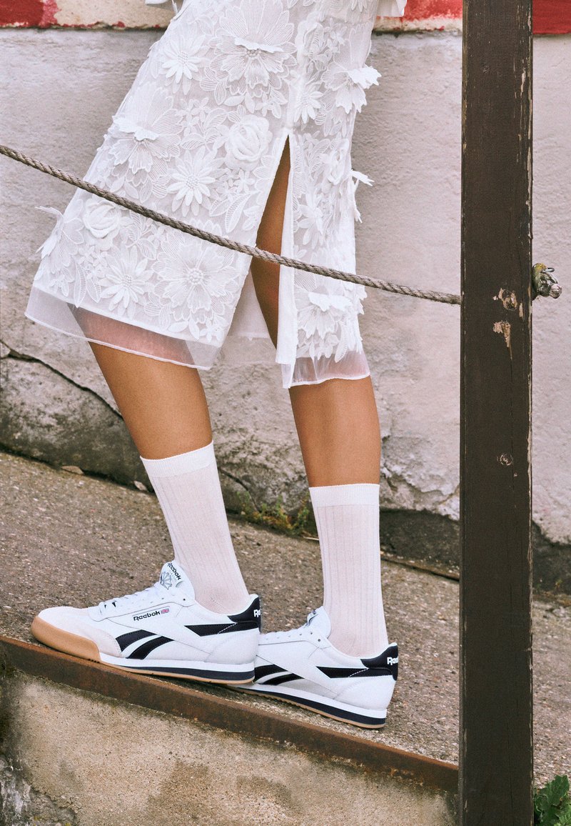 White Reebok sneakers with black accents, paired with white ribbed socks, and a white sheer floral dress with slits, on a stone surface.
