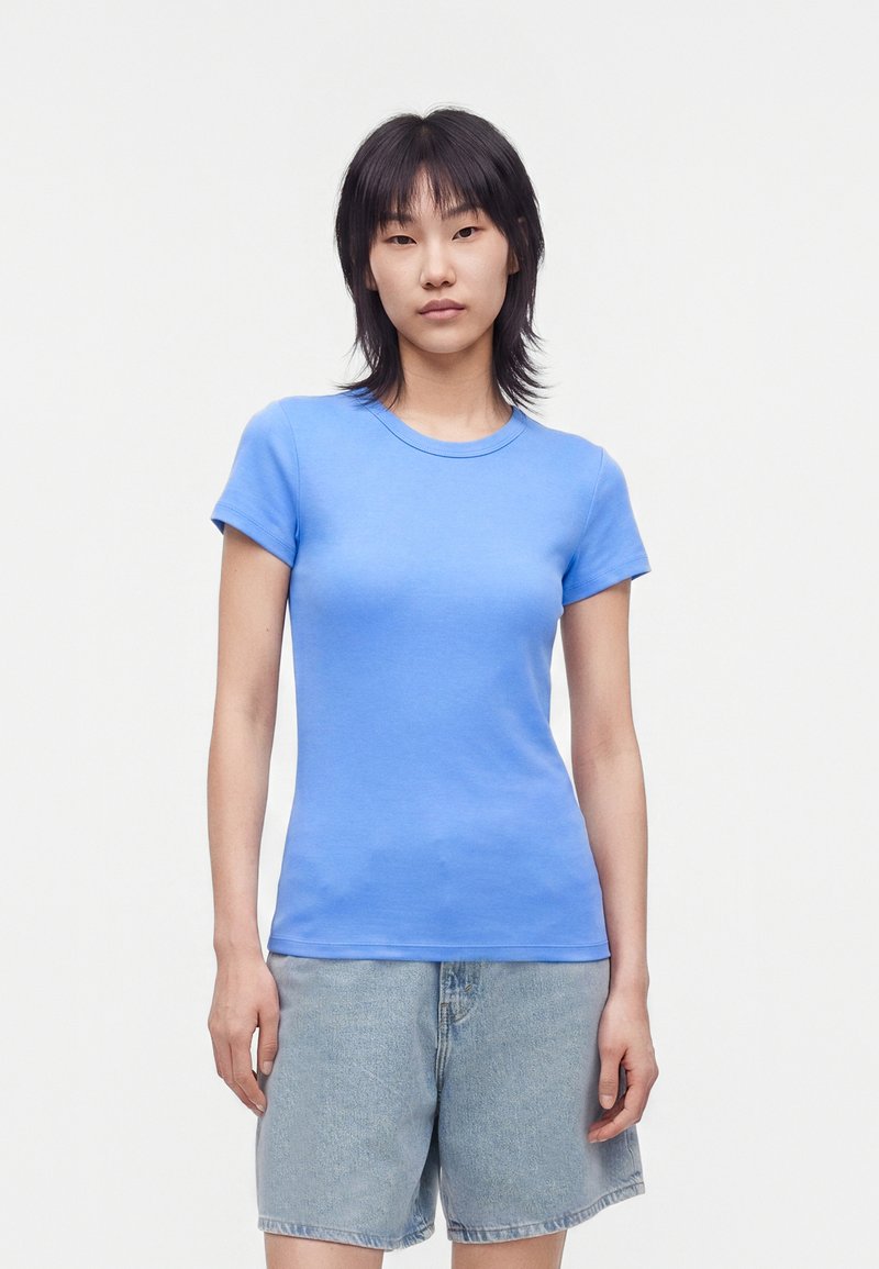 Jeune femme aux cheveux noirs et raides, portant un t-shirt bleu clair ajusté et un short en jean clair et ample, debout devant un fond uni.