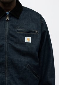 Σκούρο τζιν μπουφάν Carhartt με μαύρο γιακά, μπροστινό φερμουάρ, φερμουάρ σε τσέπη στο στήθος και γωνιακή κάτω τσέπη, φορεμένο από άτομο.