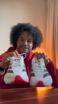 Scarpe sportive bianche con strisce nere, tomaia in mesh strutturato, dettagli rossi sul tallone e design con lacci. Presentate da una persona che le tiene in mano.