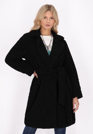 Donna bionda che indossa un cappotto nero testurizzato con cintura, top bianco, collane colorate a strati e jeans blu su uno sfondo bianco semplice.