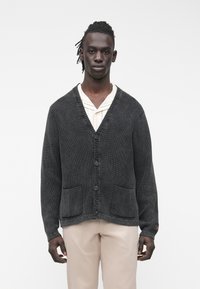 Tamsiai pilkas megztas kardiganas su V formos iškirpte, priekiniais užtrauktukais, ribiniu tekstūra ir dviem priekinėmis kišenėmis. Dėvimas ant šviesios marškinių ir smėlio spalvos kelnių.