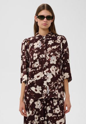 Femme aux longs cheveux bruns portant des lunettes de soleil foncées et une chemise ample foncée à motif fleuri avec un pantalon assorti, debout devant un fond uni.