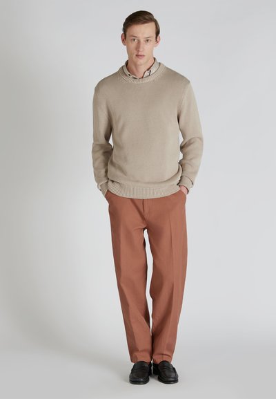 Beżowy sweter z prążkowanej dzianiny z wysokim kołnierzem, w połączeniu z rudymi spodniami i czarnymi butami. Model stoi z dłońmi w kieszeniach.