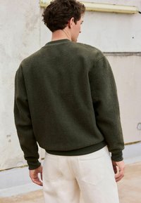 Fleece-Sweatshirt in Dunkelgrün mit einer strukturierten, weichen Oberfläche. Verfügt über einen gerippten Kragen und Bündchen, kombiniert mit hellen Hosen.