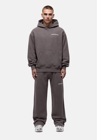 Szary, oversize'owy hoodie z przednią kieszenią i białym napisem, w komplecie z dopasowanymi dresami. Tkanina wydaje się miękka i teksturowana.