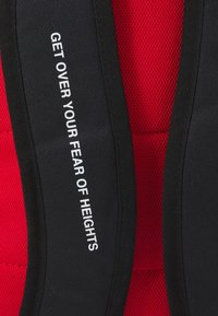 Svart och röd tygsele med en strukturerad nättextur, med texten "ÖVERVINN DIN RÄDSLA FÖR HÖJDER" tydligt visad i vitt.