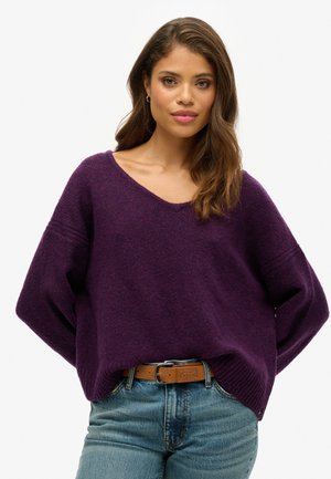Purple oversized sweater with a V-neck and ribbed hems, paired with blue denim jeans and a brown belt. Soft texture and relaxed fit.