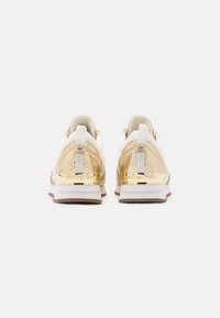 Las zapatillas blancas y doradas cuentan con un talón dorado texturizado, un acento metálico y una suela blanca con tracción marrón claro.