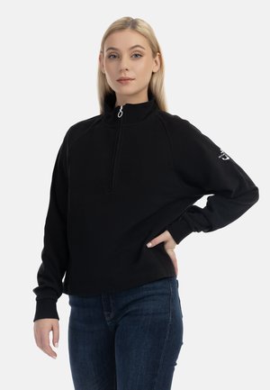 Junge Frau mit blondem Haar, die einen schwarzen Sweatshirt mit Viertel-Reißverschluss und blaue Jeans trägt, steht mit einer Hand in der Hüfte, einfarbiger Hintergrund.
