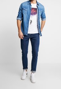 Hellblaues Denimhemd, das über einem weißen T-Shirt mit Grafikdruck getragen wird, kombiniert mit dunkelblauen Jeans und weißen Sneakers mit schwarzen Akzenten.