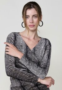 Grau mit Schlangenledermuster bedrucktes Langarmoberteil mit V-Ausschnitt. Das Model trägt große schwarze Creolen, die Haare sind leicht wellig, vor einem hellen Hintergrund präsentiert.