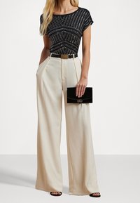 Žena oblékající černý vzorovaný top s krátkým rukávem, krémové kalhoty s vysokým pasem a širokými nohavicemi, černý opasek, černou kabelku a černé sandály na otevřenou špičku.