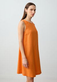 Frau mit glattem braunem Haar trägt ein ärmelloses oranges Kleid und goldene Tropfohrringe, steht vor einem schlichten hellen Hintergrund.