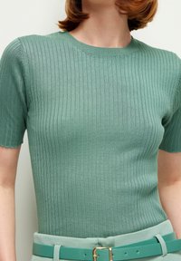 Femme portant un haut en maille côtelée vert à manches courtes, rentré dans un pantalon avec une ceinture assortie teal à boucle dorée.