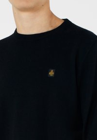 Maglione nero a maglia con scollo rotondo e polsini a coste, dotato di una piccola etichetta logo dorata sul lato sinistro del petto. Morbido al tatto.