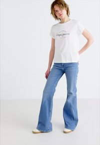 T-shirt en coton blanc avec texte de logo, associé à un jean flare bleu clair. Le mannequin se tient sur un fond neutre, portant des chaussures décontractées.