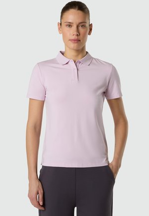 Donna in piedi con le mani lungo i fianchi, che indossa una polo a maniche corte rosa chiaro e pantaloni scuri, su uno sfondo semplice.