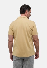 Polo à manches courtes beige, en tissu doux, avec un col classique et un ourlet droit, porté avec un pantalon à motifs gris clair.