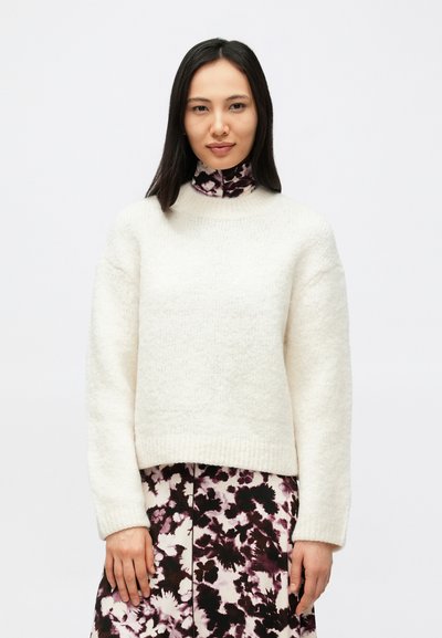 Weißer, strukturierter Pullover mit hohem Kragen und überschnittenen Schultern, getragen über einem lila Kleid mit Blumenmuster. Weiches Strickmaterial.