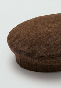 Șapcă plată maro din lână, cu un model subtil în formă de chevron, vârf rotunjit și vizieră scurtă și rigidă, expusă pe fundal alb.