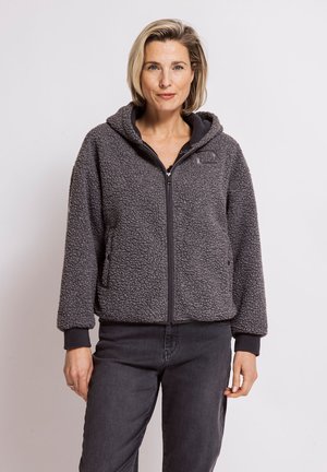 Woman with short blond hair wearing a dark gray zip-up sherpa jacket and black pants, standing against a plain light background.