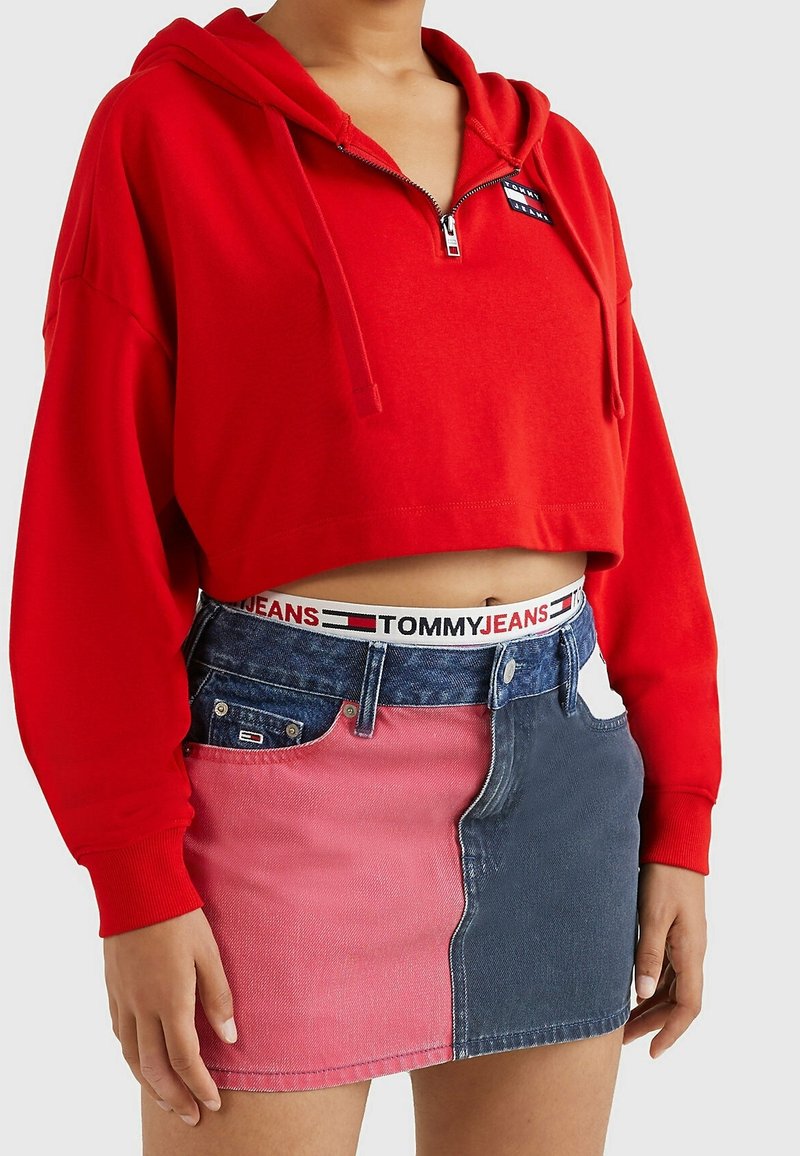 Personne portant un sweat à capuche court rouge avec une fermeture éclair à l’avant et une jupe en denim bicolore rose et bleu foncé avec une ceinture Tommy Jeans.