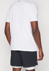 Bílý tričko s krátkým rukávem a kulatým výstřihem, na zadní straně s jemným logem, v kombinaci s černými sportovními šortkami se perforovanými detaily.