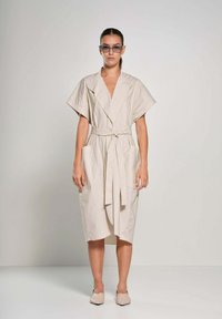 Abito avvolgente beige con maniche corte, cintato in vita, presenta due tasche anteriori e un orlo alla lunghezza del ginocchio. Tessuto di cotone morbido.