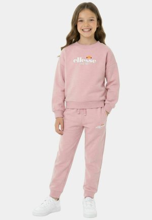 Jeune fille souriante, portant un sweat-shirt ellesse rose et un pantalon de survêtement assorti, avec des baskets blanches, debout devant un fond uni.