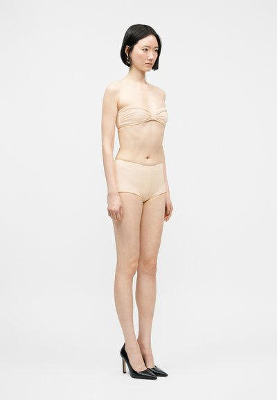 Νεαρή γυναίκα με κοντά μαύρα μαλλιά, φοράει γυμνό τοπ bandeau, ταιριαστά σορτς και μαύρες ψηλοτάκουνες γόβες, στέκεται μπροστά σε απλό ανοιχτό φόντο.