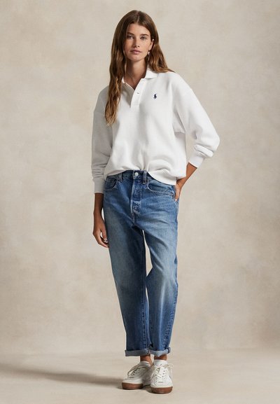 Noor naine kannab valget pika varrukaga polosärki, siniseid üleskeeratud teksapükse ja valgeid tenniseid, seistes ühevärvilise tausta ees.