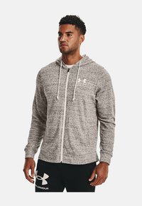 Grå zip-up hættetrøje med en tekstureret stof, hvide snore og et lille hvidt logo på venstre bryst. Stylet med sorte bukser.