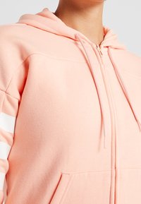 Felpa rosa con zip, dotata di cappuccio regolabile con coulisse, morbida al tatto e con strisce bianche sulle maniche. Presenta una tasca frontale e polsini a costine.