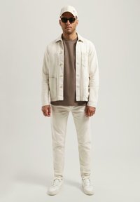 Lysfarget lin skjorte med knapper, brun crew neck skjorte under, beige bukser og hvite sneakers; accessorert med caps og solbriller.