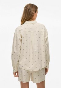 Camisa beige con sutiles patrones de diamantes y puntos, de manga larga, corte holgado y cierre de un solo botón en los puños, combinada con pantalones cortos a juego.