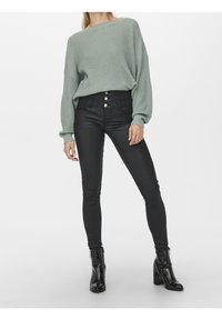 Femme portant un pull en tricot vert sauge, un pantalon skinny noir taille haute avec des boutons, et des bottines noires en cuir verni debout.