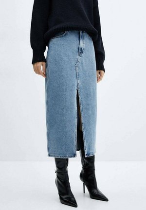 Person iført en sort sweater, lysblå midi denimnederdel med frontslids, og sorte spidse højhælede støvler, der står på et hvidt gulv.