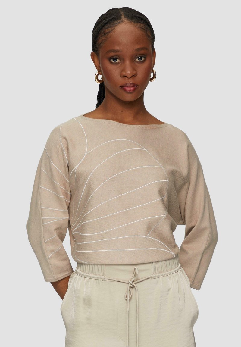 Beżowy sweter z rękawami 3/4, zdobiony białymi wzorami w paski. Miękka tekstura, okrągły dekolt, w połączeniu z jasnobeżowymi spodniami o elastycznym pasie.