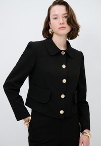Giacca nera corta con colletto arrotondato, bottoni decorativi dorati, due tasche frontali e una superficie liscia. Indossata sopra un completo coordinato.